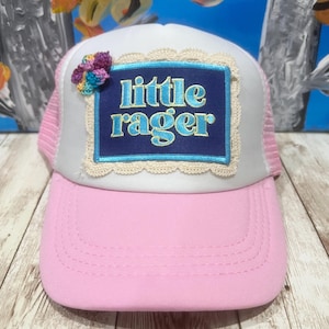 May include: A pink and white trucker hat with a pink brim and mesh back. The front features a navy blue patch with the words "little rager" in light blue, framed by a cream-colored lace trim. A small, colorful crocheted flower is on the side.