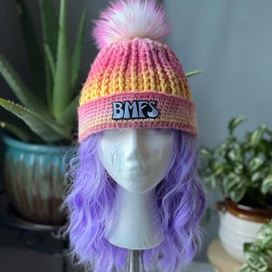 May include: A colorful knit beanie with a pink and yellow gradient, topped with a fluffy pink and white pom-pom. The hat features the letters "BMFS" on a patch. The beanie is displayed on a mannequin head with purple hair.