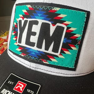May include: A white and black baseball cap with a rectangular patch. The patch features the word "YEM" in bold black letters over a turquoise background with a colorful geometric design. The cap has a black brim.