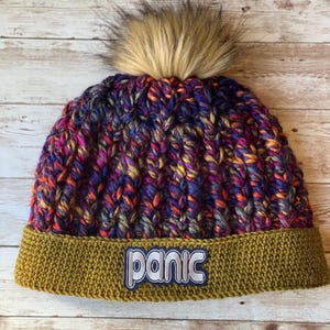May include: A colorful knit beanie with a faux fur pom-pom. The hat features a ribbed knit pattern in shades of purple, orange, and yellow. The brim is olive green with the word "panic" embroidered on it.