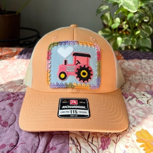 May include: A peach-coloured trucker hat featuring a square patch with a light blue background, a pink tractor, and a white heart. The patch is bordered by multi-coloured crochet stitching. A black Richardson 115 Low-Pro sticker is on the brim.