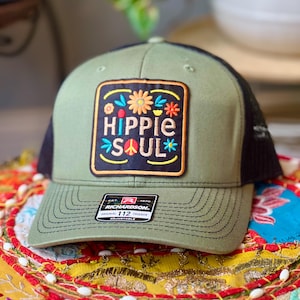 May include: Olive green trucker hat with a black mesh back and a curved brim. The front features a patch with the words "HIPPIE SOUL" in colorful letters, surrounded by floral designs. The hat is adjustable.