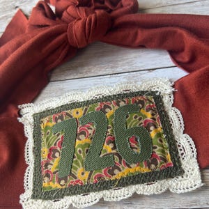 May include: A brown fabric headband with a white lace trim and a green fabric patch with the number "726" in white.