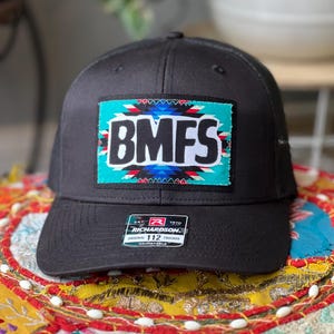 May include: Black trucker hat with a rectangular patch featuring the letters "BMFS" in white, set against a turquoise background with a Southwestern design. The hat has a curved brim and an adjustable strap. The Richardson brand label is visible.