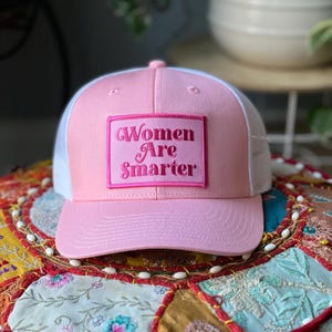 May include: A pink and white trucker hat with the text "Women Are Smarter" embroidered on a pink patch. The hat has a curved brim and a mesh back. The hat is sitting on a colorful, patterned surface.