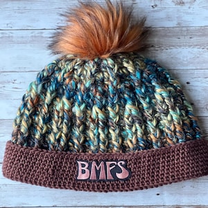 May include: A handmade knit beanie in shades of blue, brown, and yellow with a brown brim. The beanie features a fluffy orange pom-pom on top and the letters "BMFS" embroidered on the brim.
