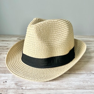 May include: A beige straw fedora hat with a black ribbon band. The hat is sitting on a light wood surface.