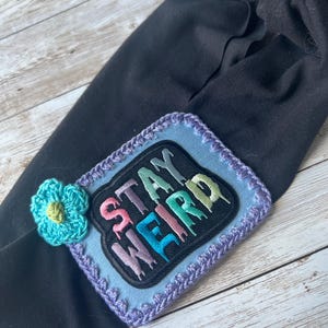 May include: A black fabric headband with a blue crocheted border and a colorful "Stay Weird" patch. A small blue crocheted flower is attached to the headband.
