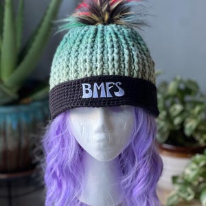 May include: A handmade knit beanie with a gradient teal and green color scheme, a black band with the letters "BMFS", and a colorful faux fur pom-pom. The hat is displayed on a mannequin head with purple hair.