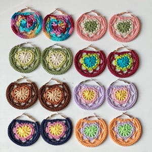 May include: A collection of colorful, handmade crochet hoop earrings. The earrings feature various color combinations, including pink, green, blue, and orange, with a heart-shaped design in the center. The earrings are displayed on a white surface.