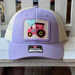 May include: A lavender trucker hat with a cream-colored mesh back and a lavender bill. The front features a patch with a pink tractor and a blue heart. The hat has a Richardson 115 Trucker tag.