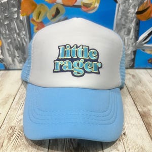 May include: A light blue and white trucker hat with the text "Little Rager" in turquoise. The hat has a light blue brim and a white front panel with a mesh back.