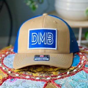 May include: A tan and blue trucker hat with a blue brim and mesh back. The front features a blue rectangle with the letters "DMB" in white. The hat has a Richardson 112 tag.