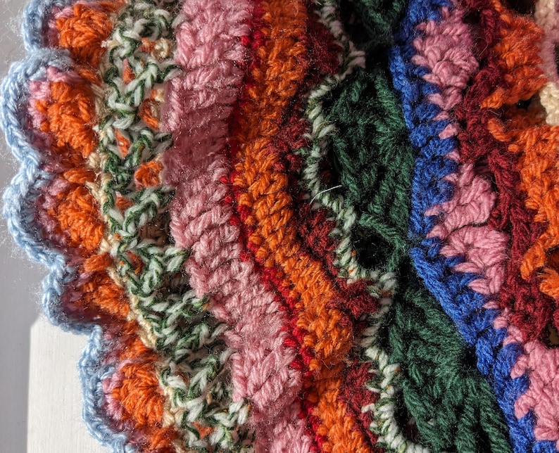May include: Close-up of a colorful, handmade crochet item. The piece features a variety of colors including orange, pink, green, blue, and red. The edges have a scalloped design. The texture is visible, showcasing the yarn's individual stitches.