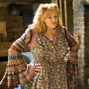 May include: A woman wearing a floral print dress with a knitted, multi-colored sweater. The sweater has bell sleeves with orange, brown, and blue stripes. The dress has a V-neck and a pocket on the front.