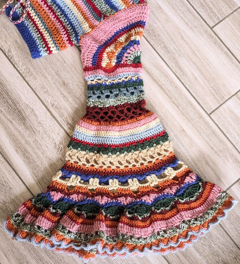 May include: A colorful, handmade crochet dress with a flared skirt and long sleeves. The dress features a variety of colors including pink, blue, orange, and green. The crochet work has a detailed, open-weave pattern, and the dress is displayed on a wooden surface.