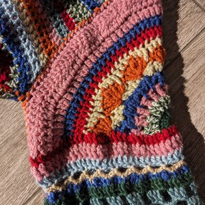 May include: Close-up of a colorful, handmade crochet item. The piece features a variety of colors including pink, blue, orange, red, and green. The texture is thick and the stitches are clearly visible, suggesting a cozy, handcrafted textile.