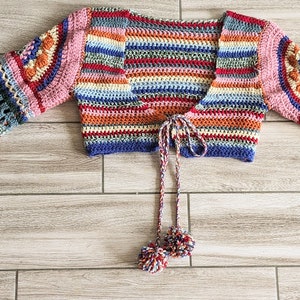 May include: A colorful, handmade crochet top with flared sleeves. The top features a variety of colors including pink, blue, orange, and green. The sleeves have a detailed, openwork design. The top has a tie closure with two small pom-poms.