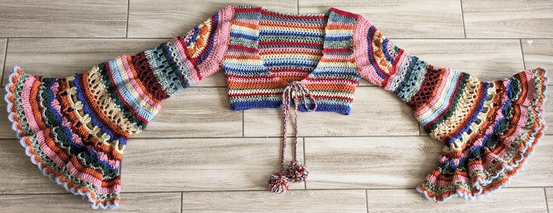 May include: A colorful, handmade crochet top with flared sleeves. The top features a variety of colors including pink, blue, orange, and green. The sleeves have a detailed, openwork design. The top has a tie closure with two small pom-poms.