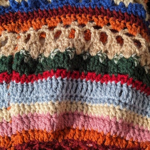 May include: Close-up of a handmade crochet item with a variety of colors. The piece features horizontal stripes in shades of orange, blue, red, green, pink, and cream. The texture is visible, showcasing the intricate stitch work.