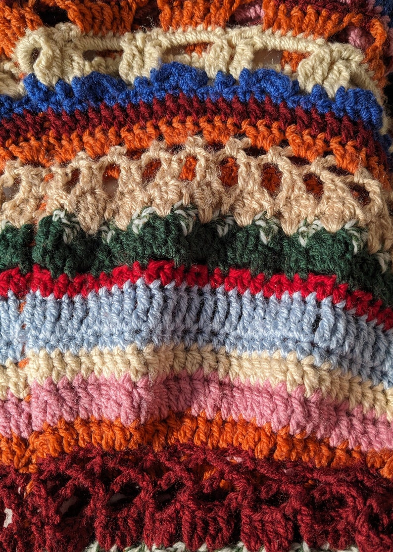 May include: Close-up of a handmade crochet item with a variety of colors. The piece features horizontal stripes in shades of orange, blue, red, green, pink, and cream. The texture is visible, showcasing the intricate stitch work.