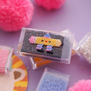May include: A small, clear plastic box filled with colorful beads. The beads are arranged in a pattern to create a cartoon image of a pencil with a smiling face. The box is sitting on a purple surface.