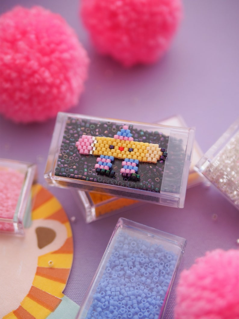 May include: A small, clear plastic box filled with colorful beads. The beads are arranged in a pattern to create a cartoon image of a pencil with a smiling face. The box is sitting on a purple surface.