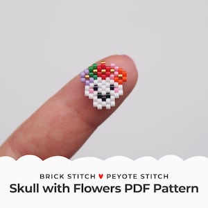 May include: A white skull bead with a colorful flower crown made with seed beads. The skull has a smiling face with black eyes and a black mouth. The flower crown is made with red, orange, yellow, green, blue, and purple beads. The bead is on a finger.