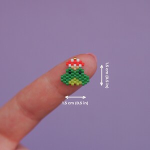 May include: A green frog with a red flower on its head, made of beads. The frog is about 0.5 inches tall.