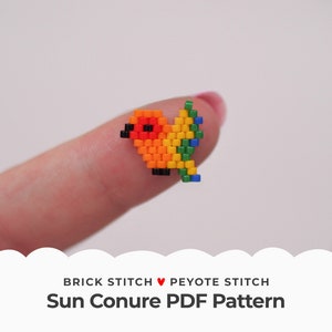 Op de afbeelding: Een kleine, gepixelde vogel gemaakt van kleurrijke kralen, voornamelijk oranje, geel, groen en blauw, met een zwarte snavel. De vogel wordt op een vinger gehouden. De tekst onderaan luidt "Sun Conure PDF Pattern".