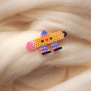 May include: A small, colorful beaded pin shaped like a cartoon character. The character is yellow with a pink nose, blue eyes, and pink and blue legs. The pin is on a white background.