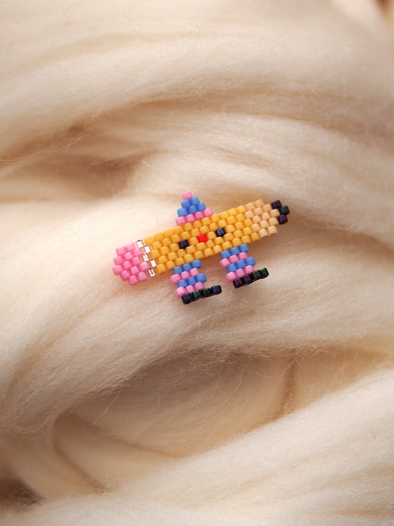 May include: A small, colorful beaded pin shaped like a cartoon character. The character is yellow with a pink nose, blue eyes, and pink and blue legs. The pin is on a white background.