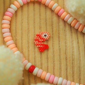 Puede incluir: Un brazalete de cuentas rosa y blanco con un amuleto de Pac-Man pixelado rojo.