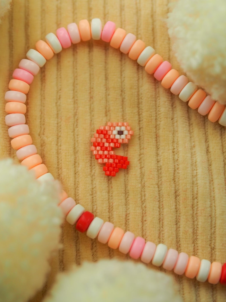 Puede incluir: Un brazalete de cuentas rosa y blanco con un amuleto de Pac-Man pixelado rojo.