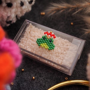 May include: A green and red beaded Christmas tree ornament with a gold bead on top. The ornament is sitting in a clear plastic container filled with white beads.
