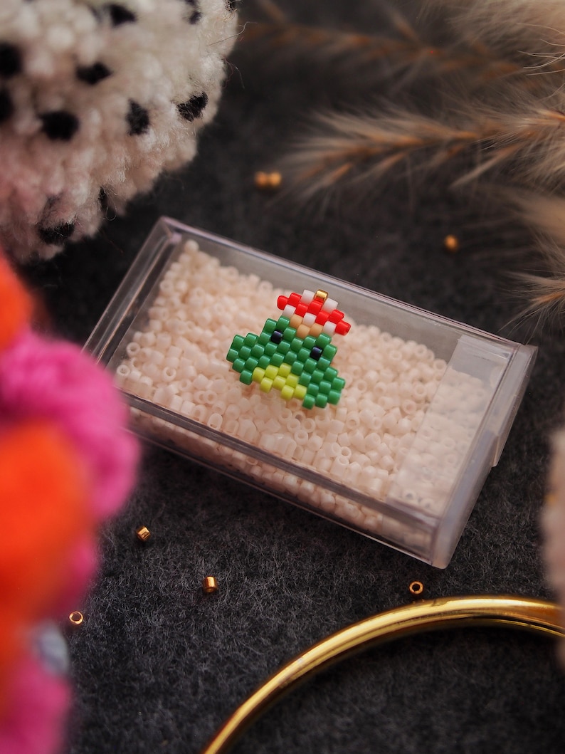 May include: A green and red beaded Christmas tree ornament with a gold bead on top. The ornament is sitting in a clear plastic container filled with white beads.