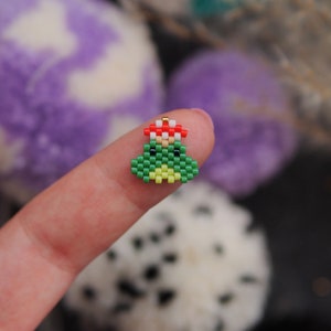 May include: A small green Christmas tree ornament made of beads with a red and white bead hat. The ornament is being held by a finger.