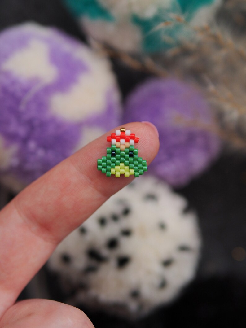 May include: A small green Christmas tree ornament made of beads with a red and white bead hat. The ornament is being held by a finger.