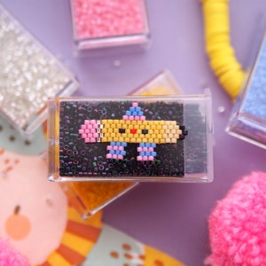 May include: A yellow, pink, and blue pixelated pencil with a smiling face, beaded on a black background. The pencil is in a clear plastic frame.