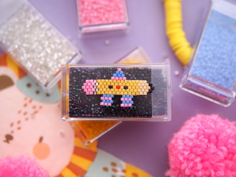 May include: A yellow, pink, and blue pixelated pencil with a smiling face, beaded on a black background. The pencil is in a clear plastic frame.