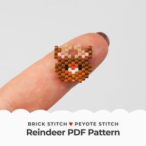 May include: A close-up of a beaded reindeer charm made with brown, beige, pink, red, and white beads. The charm is being held by a finger and is a small, detailed piece of beadwork.