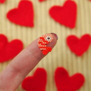 Puede incluir: Una yema del dedo con un peque&ntilde;o coraz&oacute;n de cuentas rojas y blancas sobre ella. El coraz&oacute;n est&aacute; rodeado de corazones de fieltro rojo.