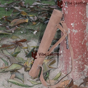 May include: A brown leather quiver with a shoulder strap, designed for arrows. The cylindrical quiver has a reinforced base and flap closure. The adjustable strap is attached with metal hardware. The background shows fallen leaves and a tree trunk.
