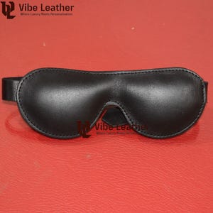 May include: Black leather eye mask with a curved design and a strap. The mask has a smooth texture and is designed to cover the eyes. The text "Vibe Leather" is visible in the upper left and lower center.