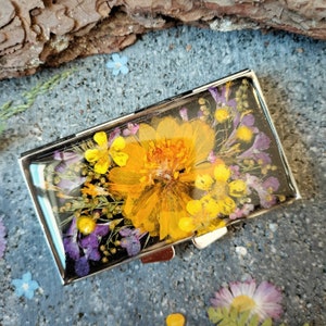 May include: A silver metal box with a clear top filled with pressed yellow and purple flowers. The flowers are arranged in a random pattern and are surrounded by green leaves.