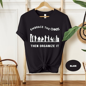 May include: Black t-shirt with white text that reads "Embrace the Chaos then Organize It". The text is surrounded by a graphic of stick figures in an office setting.