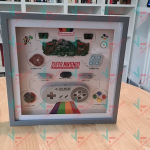 May include: A framed display of a disassembled Super Nintendo Entertainment System. The display includes the motherboard, controller buttons, and a Super Nintendo controller. The frame is grey and the background is white.