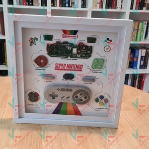 May include: A white framed shadow box featuring a Super Nintendo controller and various components from the console. The box has a white background with the text "Super Nintendo Entertainment System" and the Nintendo logo.
