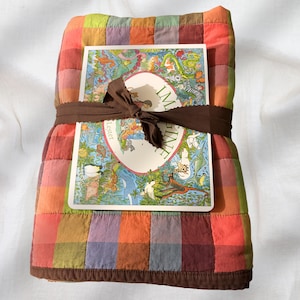 May include: A folded quilt featuring a multicoloured grid pattern in shades of orange, red, purple, green, and yellow, tied with a dark brown ribbon. A rectangular book titled IMAGINE by Lester is placed on top, decorated with colourful animal illustrations.