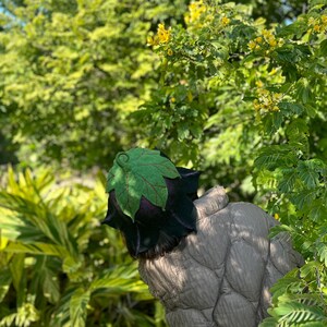 Puede incluir: Un sombrero &uacute;nico con un dise&ntilde;o de hoja. El sombrero presenta una hoja verde en la parte superior y una base de color p&uacute;rpura oscuro y negro. El sombrero est&aacute; rodeado de follaje verde y flores amarillas.